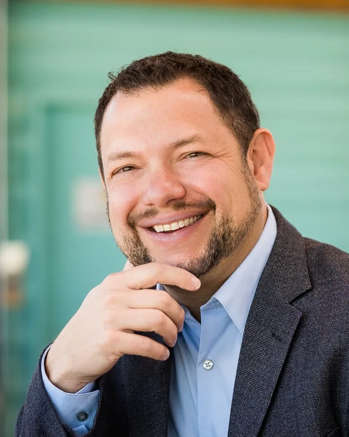 Tim Nisly, smiling, in a charcoal jacket and light blue shirt, with a hand at his chin.
